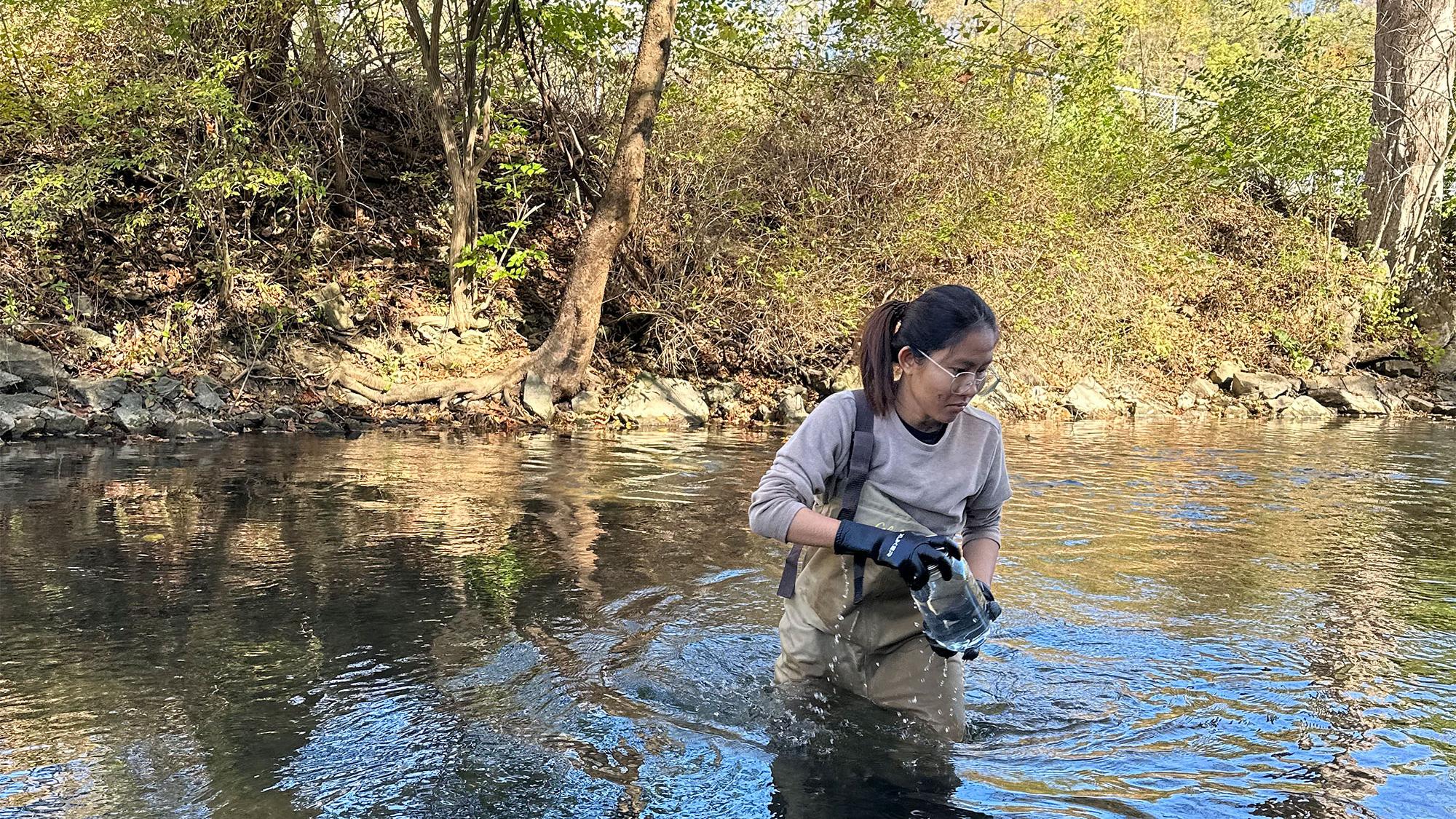 A researcher, waist deep in water, collects water and sediment samples from a river in a glass jar