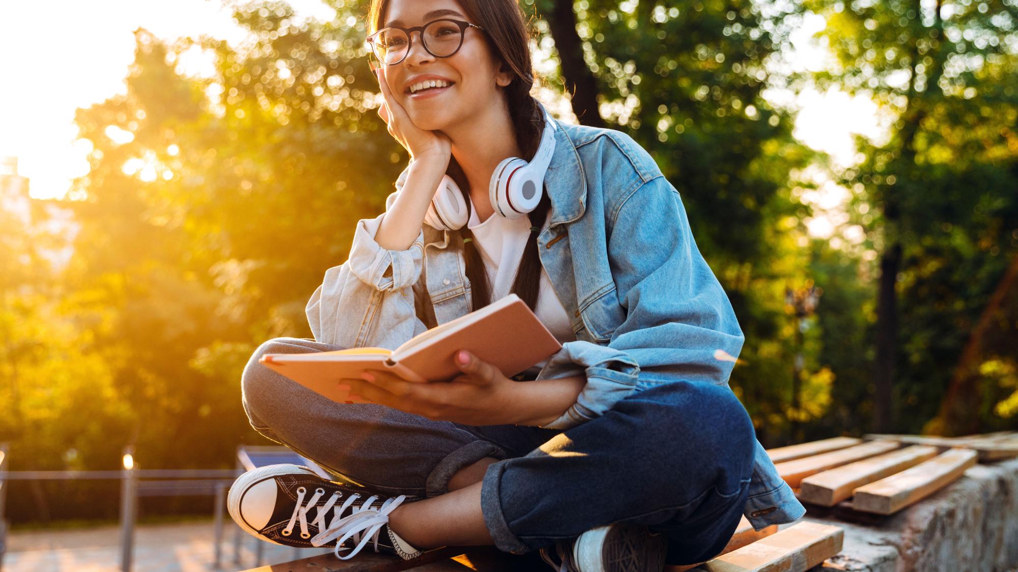 A smiling teenager sits outdoors on a bench reading a book on a sunny summer afternoon.