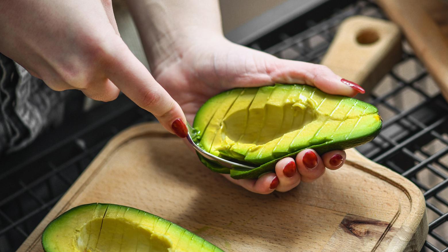 A woman's hands scoop a ripe avocado above a cutting board in a kitchen