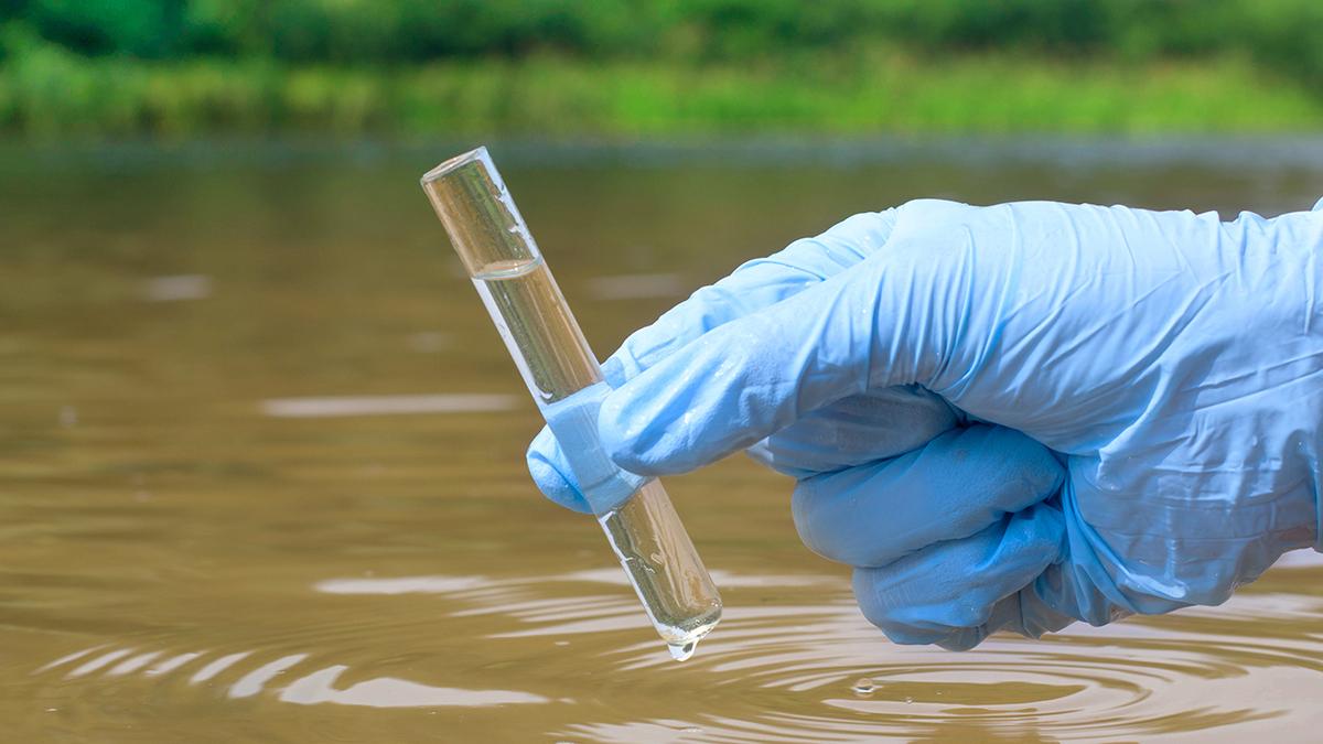 a gloved hand holds a beaker of water above a body of water