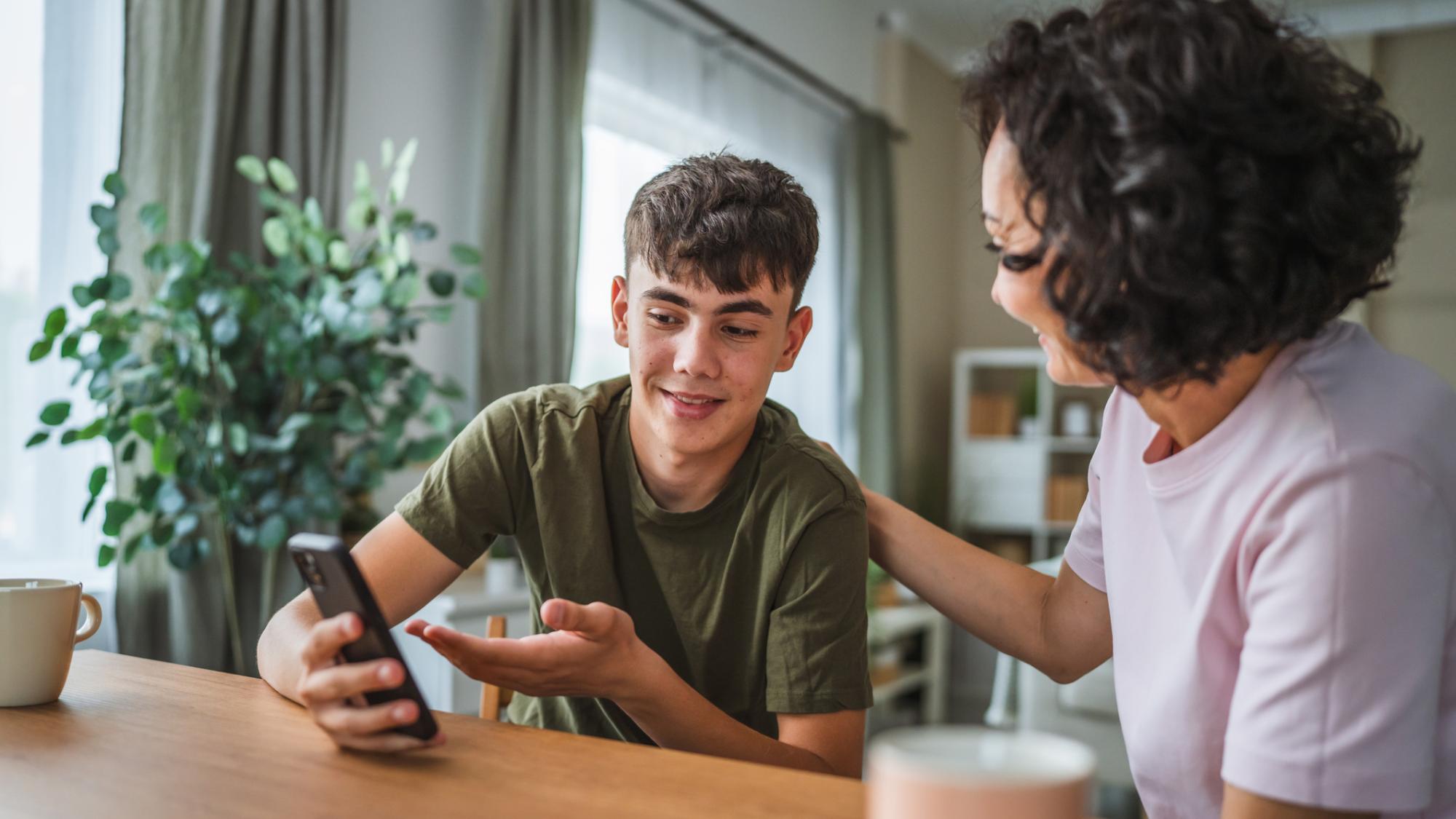 A pleased teenage boy discussing something on his telephone with his smiling mother, who rests her hand on his shoulder.