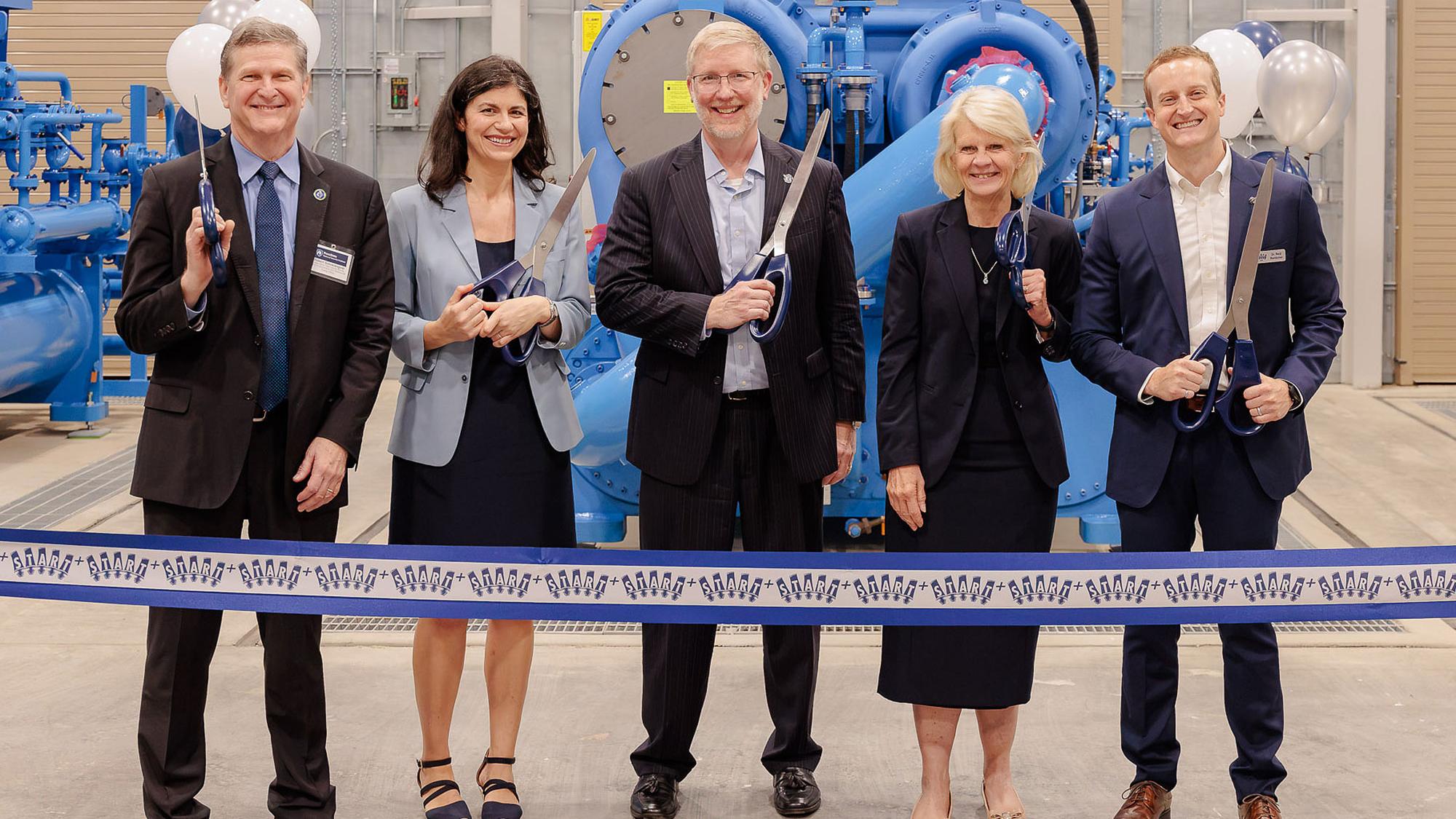 Leadership from industry, government and academia pose in front of a giant mechanical compressor, holding big scissors to cut a START+ branded ribbon