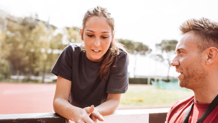 An athlete and coach, outdoors at a track, discuss something on a clipboard 