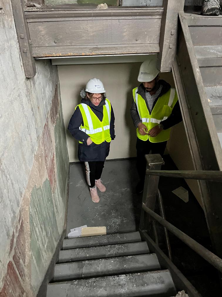 Two individuals wearing hard hats and yellow vests examine stairs in a building