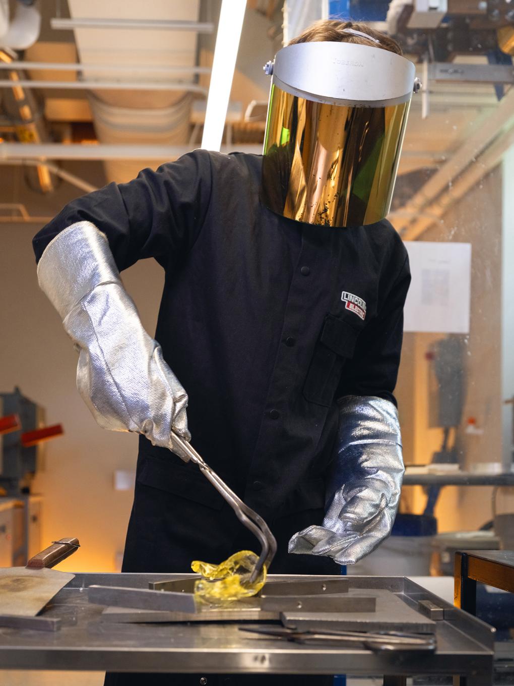 A person wearing a face heat shield molds glass from a forge