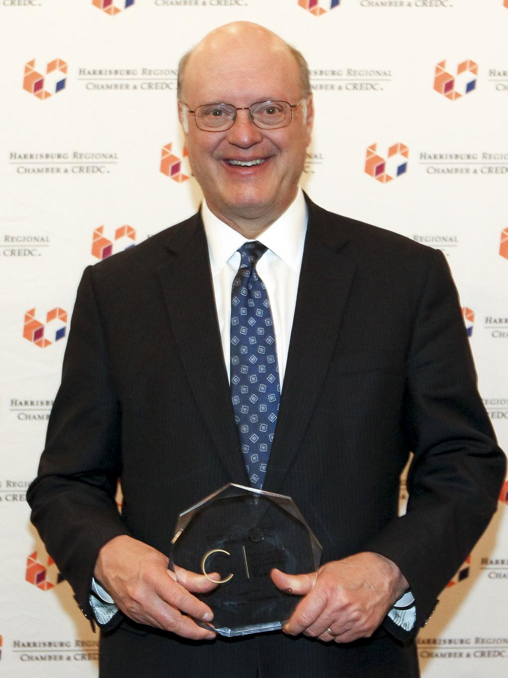 Dr. Craig Hillemeier, dressed in a suit, poses in front of a backdrop with the Harrisburg Regional Chamber and CREDC logos. He is smiling and holding an award.