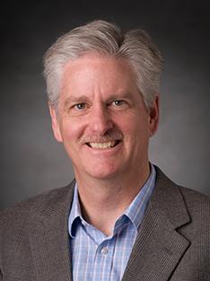 A headshot of a man in a blue button-down shirt and gray suit jacket smiling at the camera.