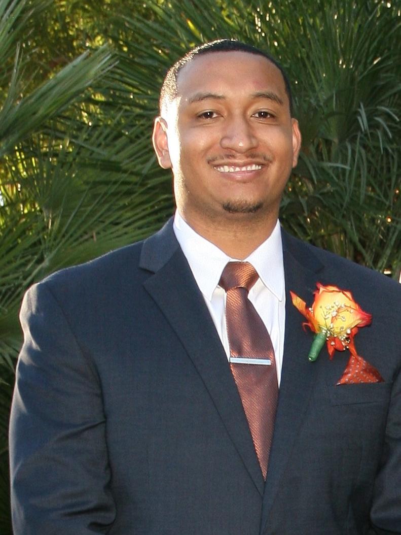 Jordan Hughey poses for a photo in a dark suit and tie and white shirt. A green tree is in the background.