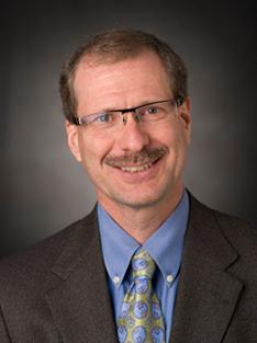 Headshot of a man with a mustache and glasses wearing a button-down shirt, tie and suit jacket 