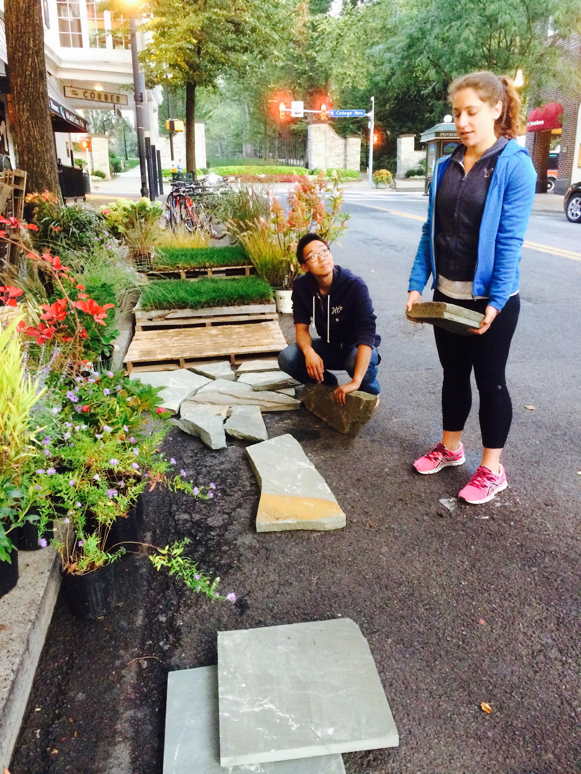 Two students working with flowers, plants, rocks, and tiles in street
