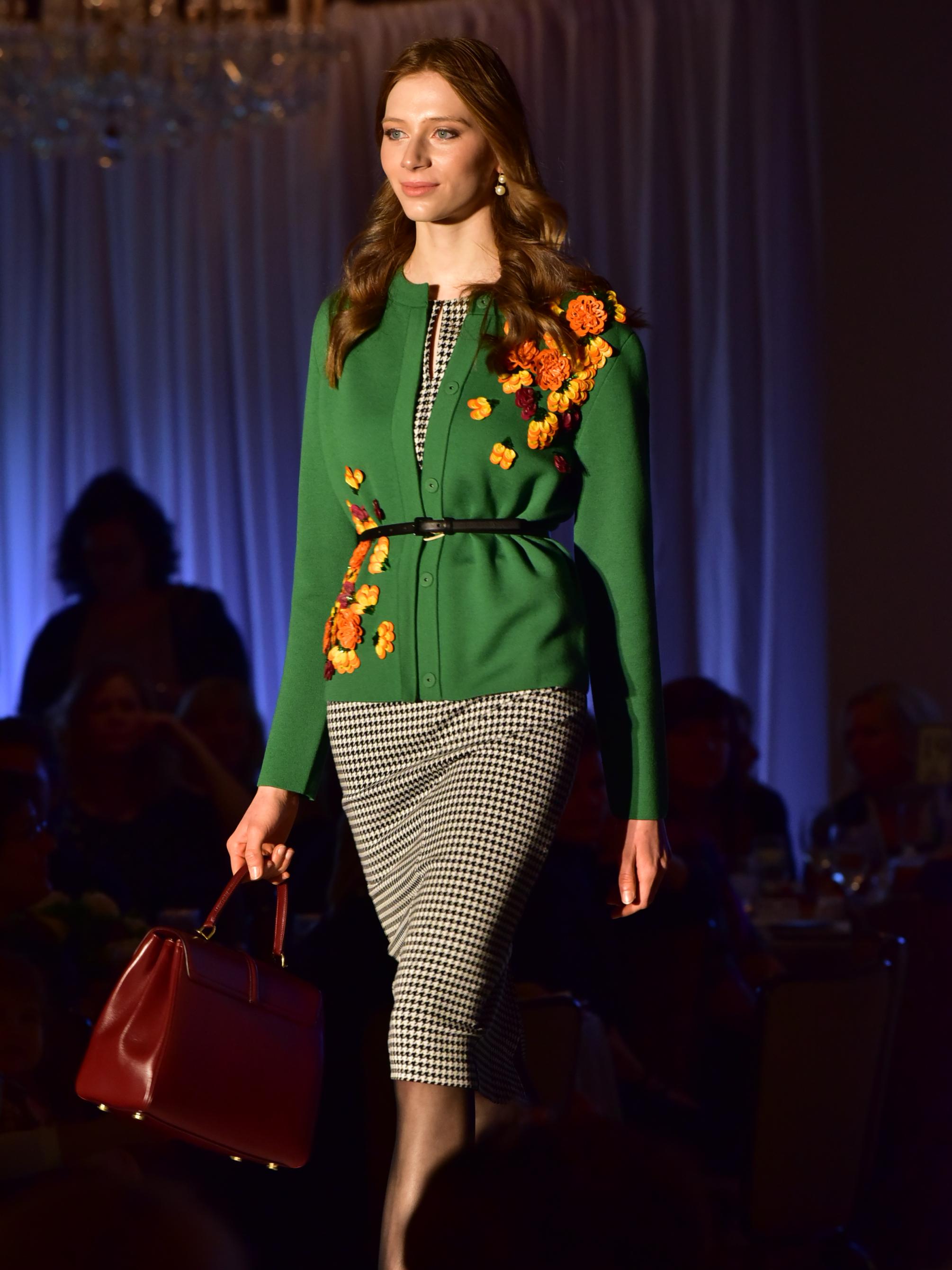 A model walks the runway wearing a checkered dress with a floral design sweater over it, and carrying a handbag in her right hand.