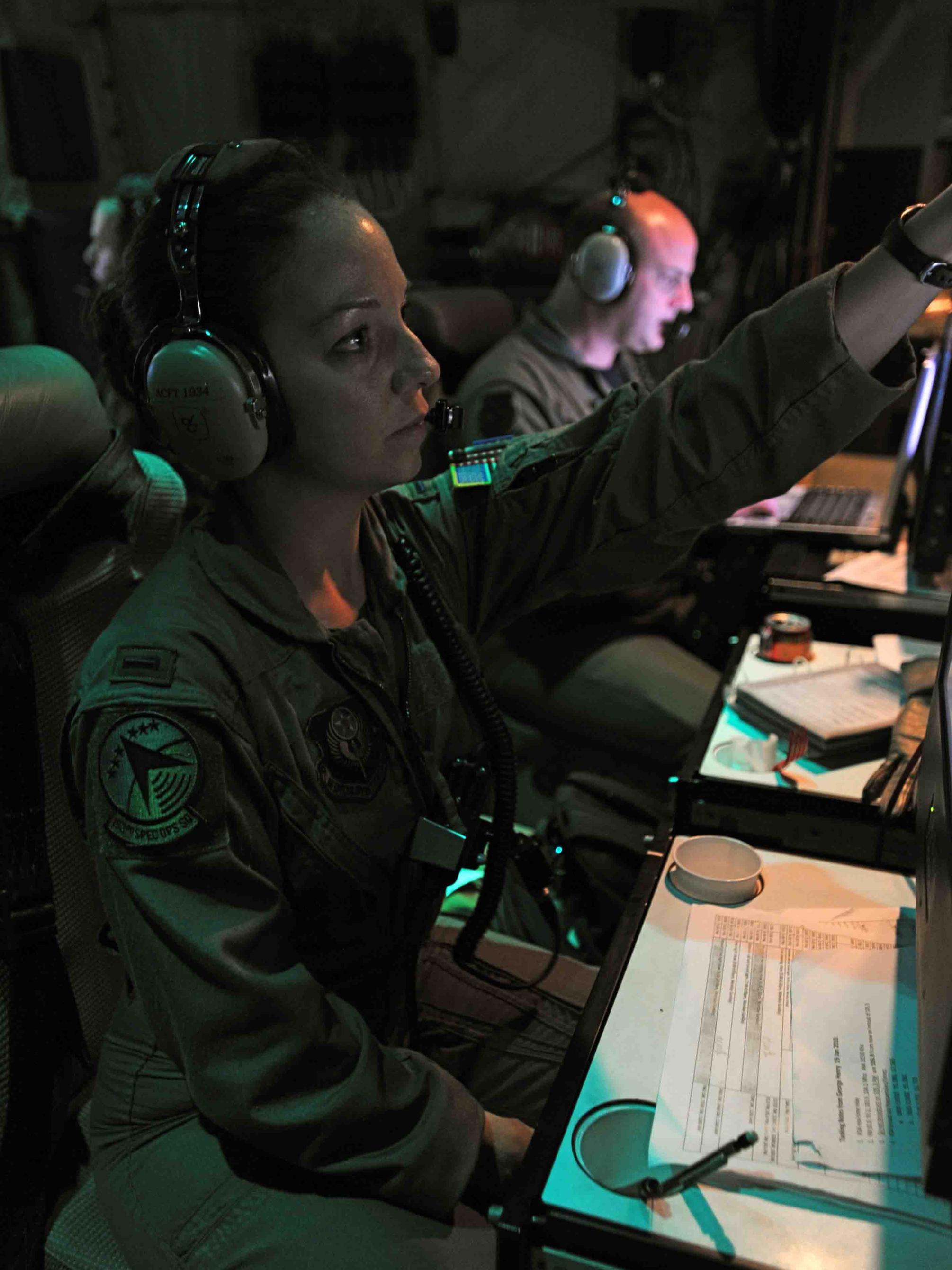 A woman in the armed forces wears headphones and points at a computer screen in a dimly lit room.