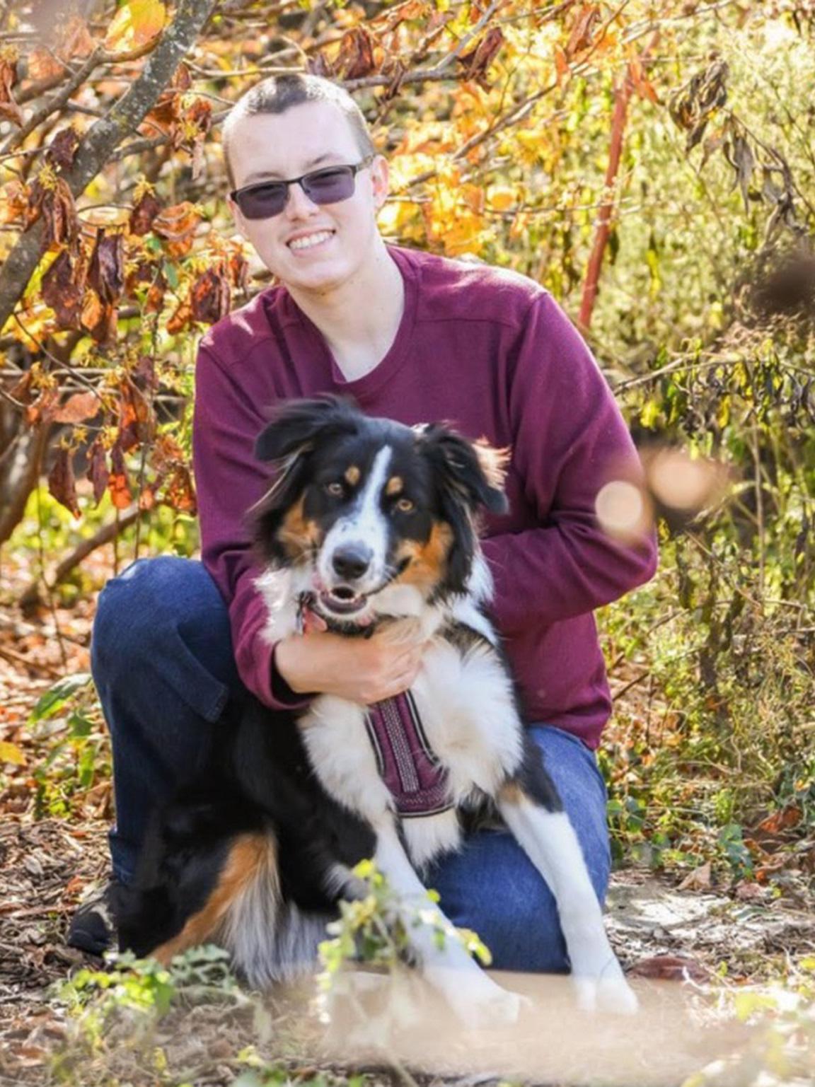 Dylan Gilbert smiles and kneels in front of a tree with his Australian shepherd, Mocha, who is brown, white and black. Dylan wears glasses, a shirt and jeans.