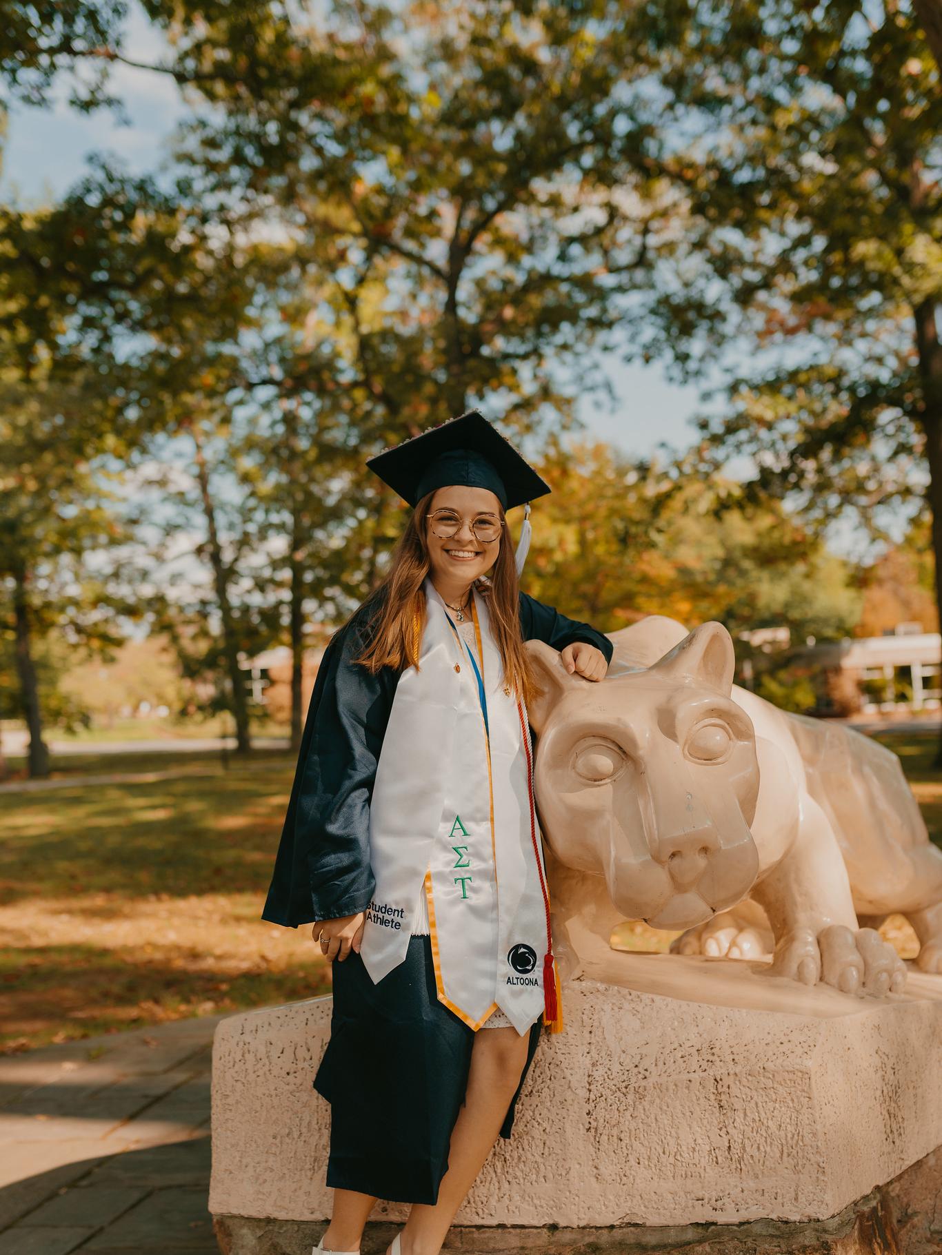 Woman poses in graduation cap and gown with a Lion Shrine statue