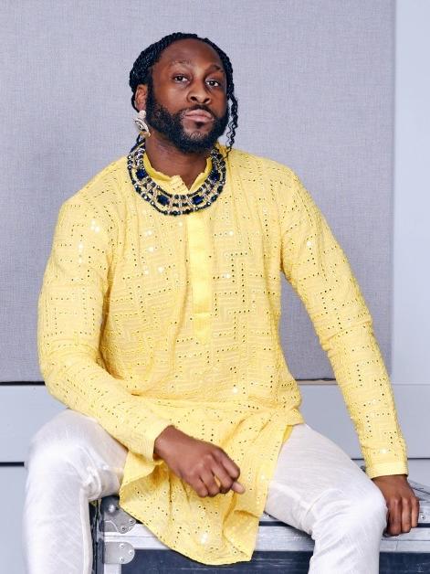 A man of color wearing braids in his hair and a flowing tunic sits on a bench.