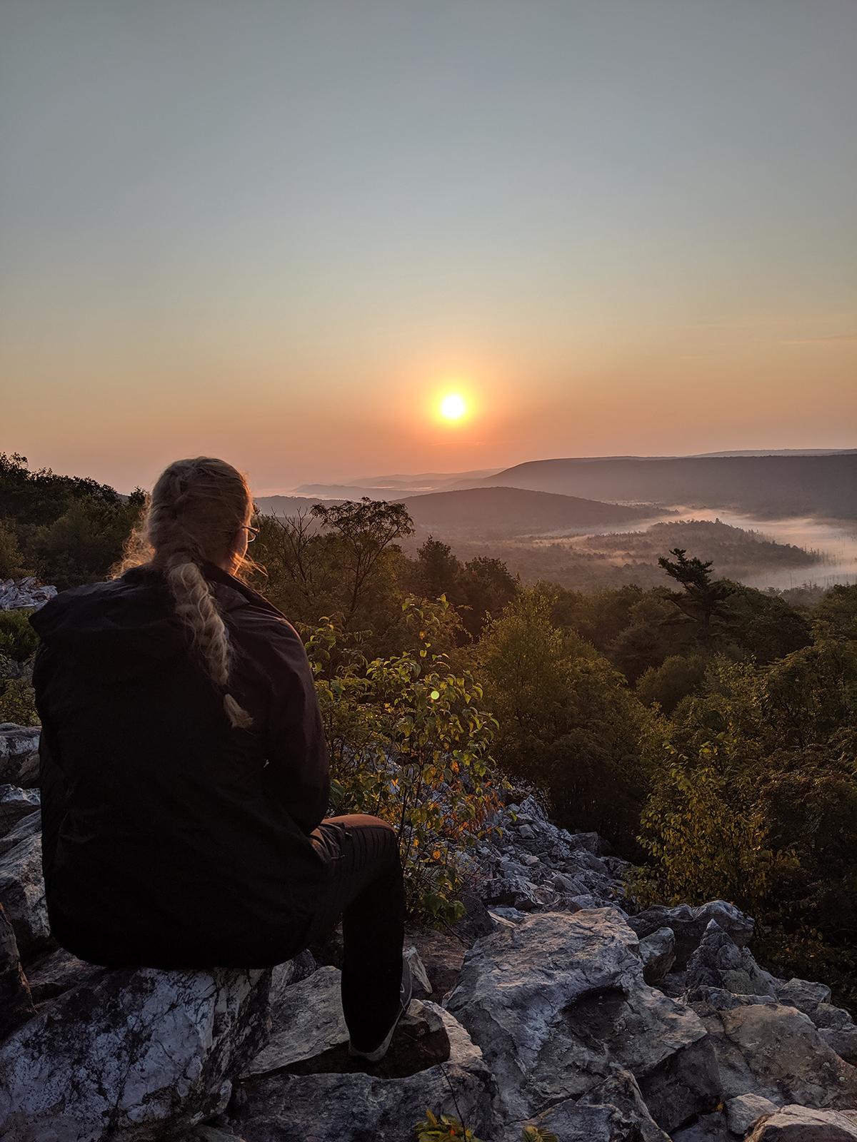 A student sitting atop a rocky ridge watches the sun rise over the distant mountains