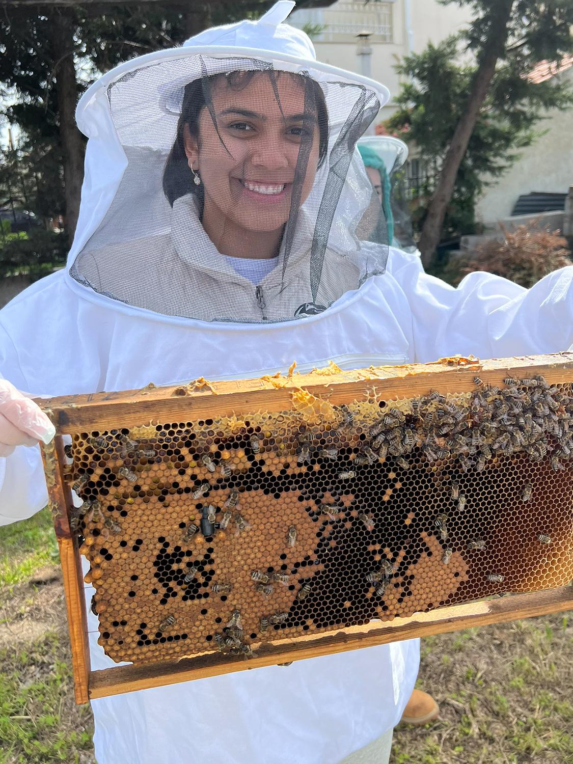 Michelle Kengkart wearing a beekeeper's suit and holding a honeycomb panel while visiting From A to Bee Farm in Pallini, Greece.