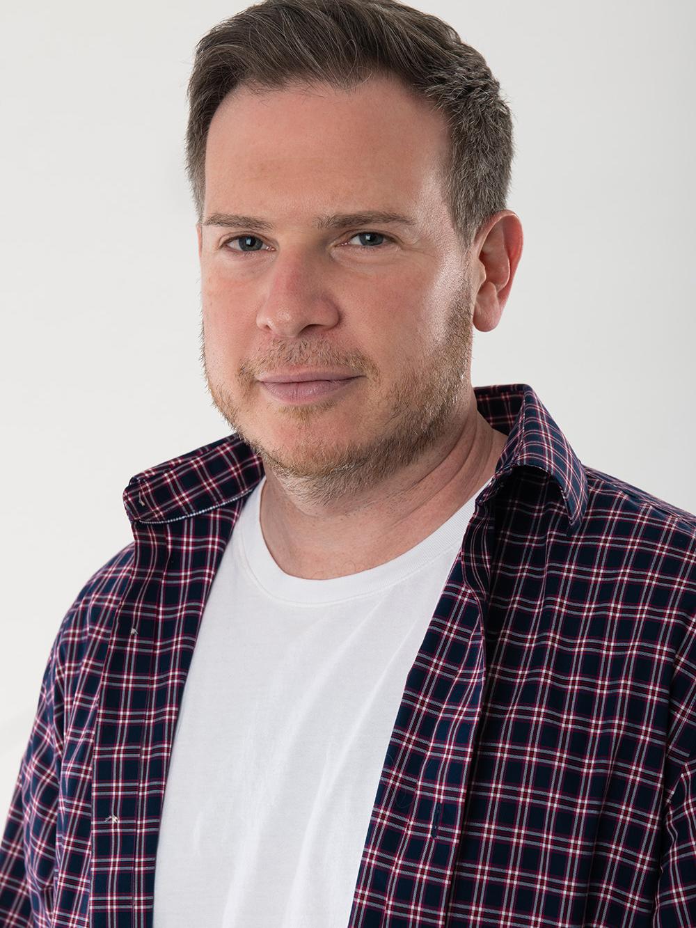 Headshot of Ira Rosensweig, wearing a white t-shirt with an unbuttoned red plaid oxford