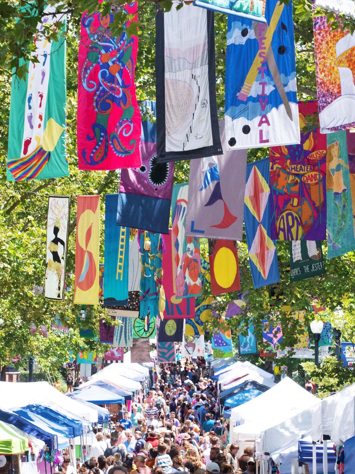 A crowd mills about on a tree-lined street, with colorful flags hanging overhead.