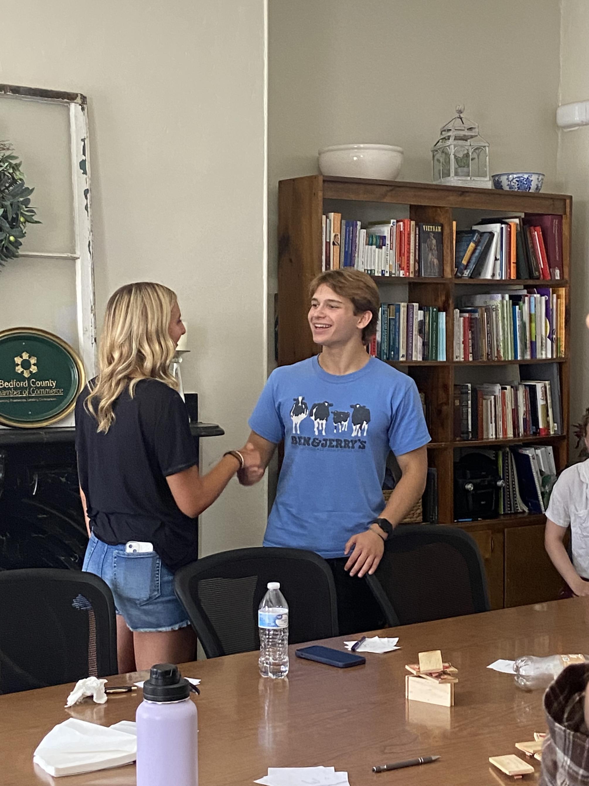 Two students shaking hands at a table in front of a book shelf.