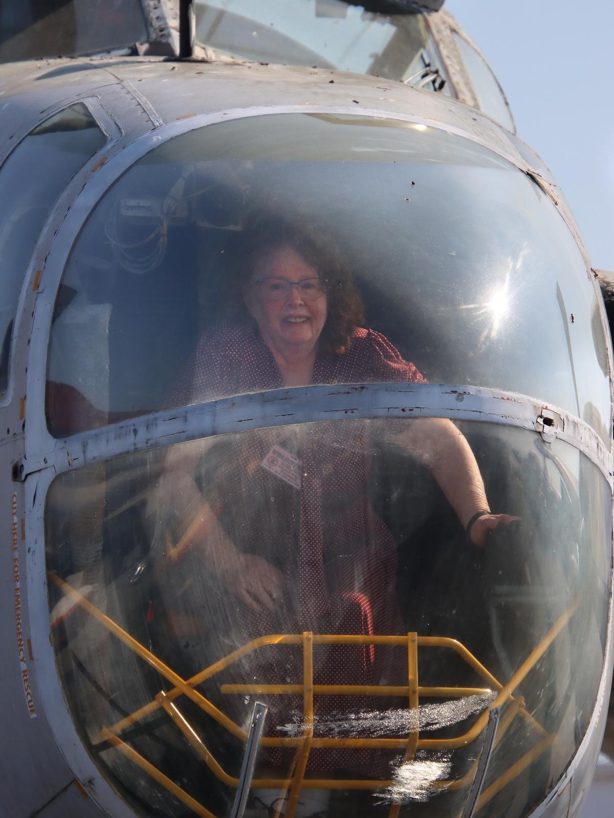 A woman sits in the cockpit of a World War II helicopter. She is wearing glasses and a name badge is clipped to her clothing. Looped wires are hanging from an instrument to the left of her head. A series of metal bars are visible at the bottom of the cockpit.