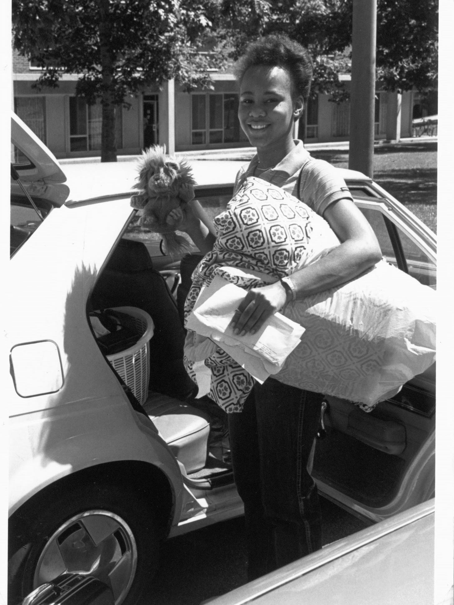 A female student standing next to a car  holding blankets and a stuffed animal