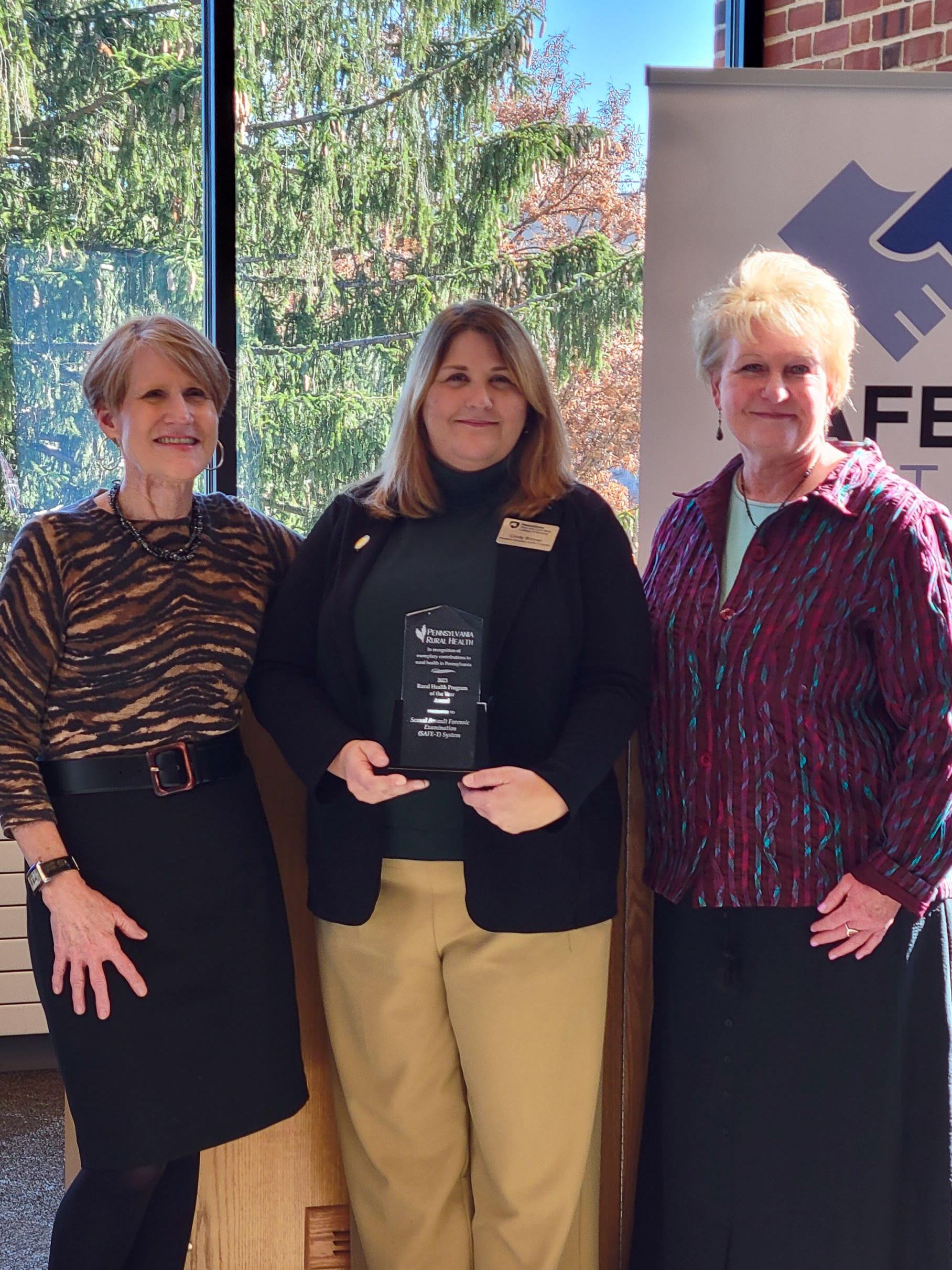 Cynthia Bittner, holding award, poses with Lisa Davis and Karen Burke in front of large glass windows.