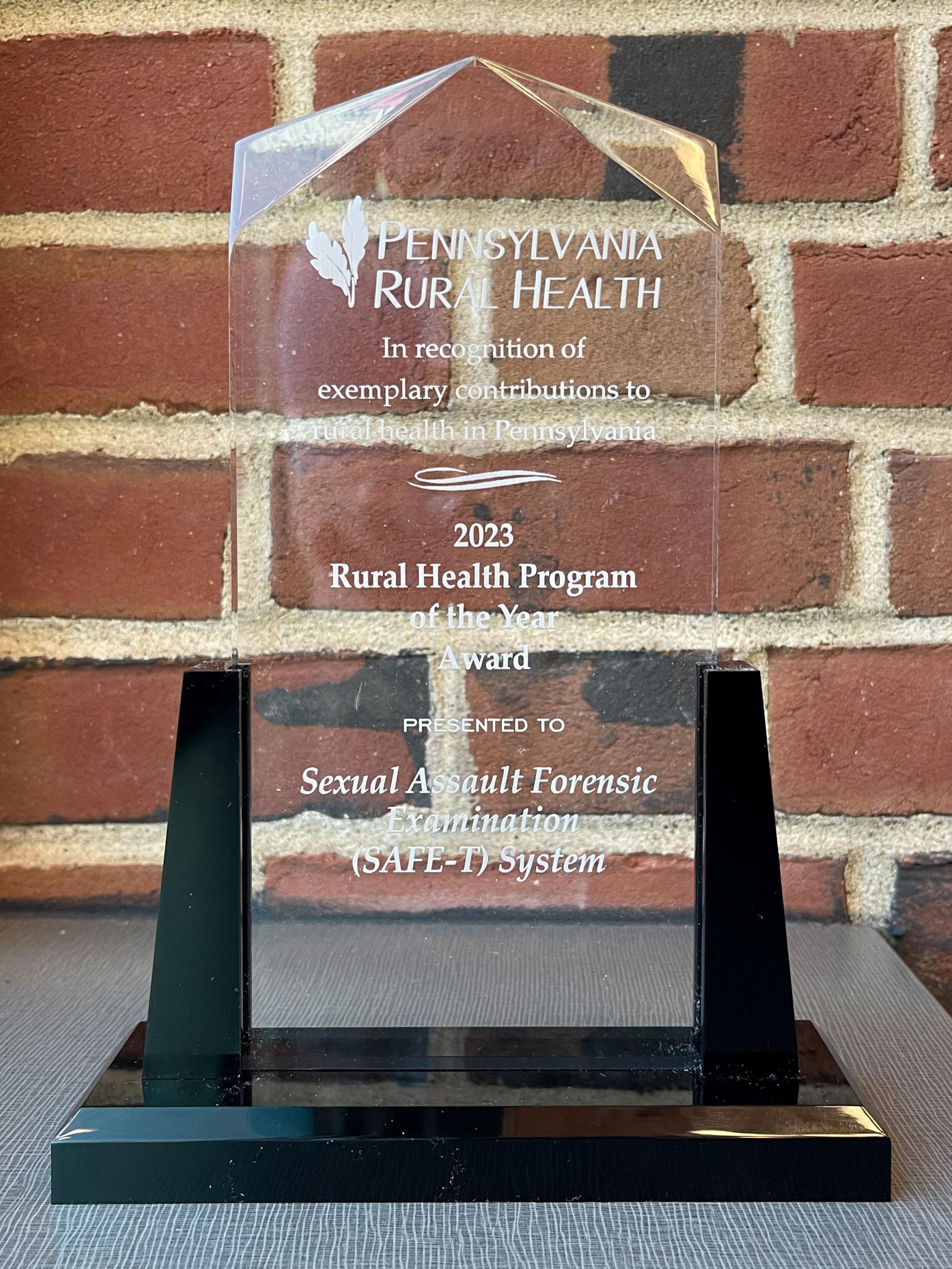 Glass plaque against a brick background that says "Pennsylvania Rural Health in recognition of exemplary contributions to rural health in Pennsylvania, 2023 Rural Health Program of the Yeard Award presented to Sexual Assault Forensic Examination (SAFE-T) System."