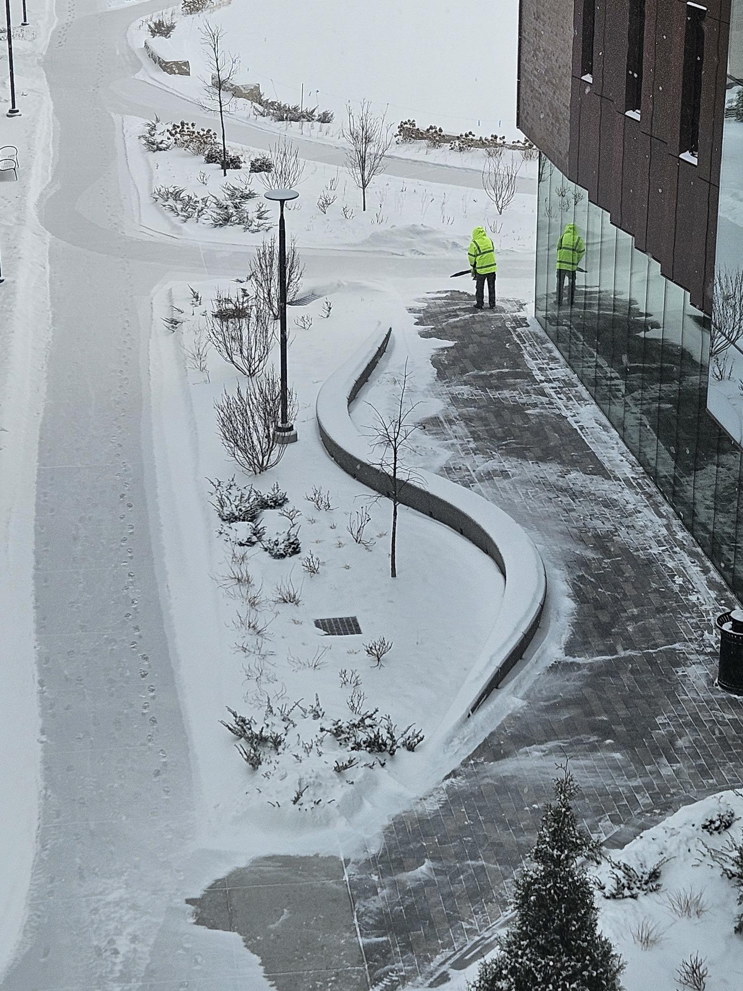 An employee wearing a yellow coat clears snow from a sidewalk