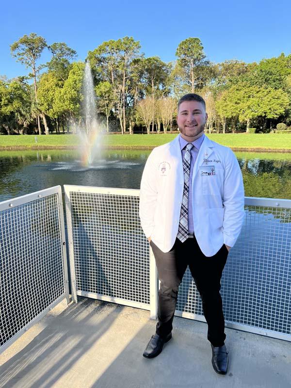 Man in white doctor's jacket standing on balcony in front of a pond with a water fountain gushing from inside it.