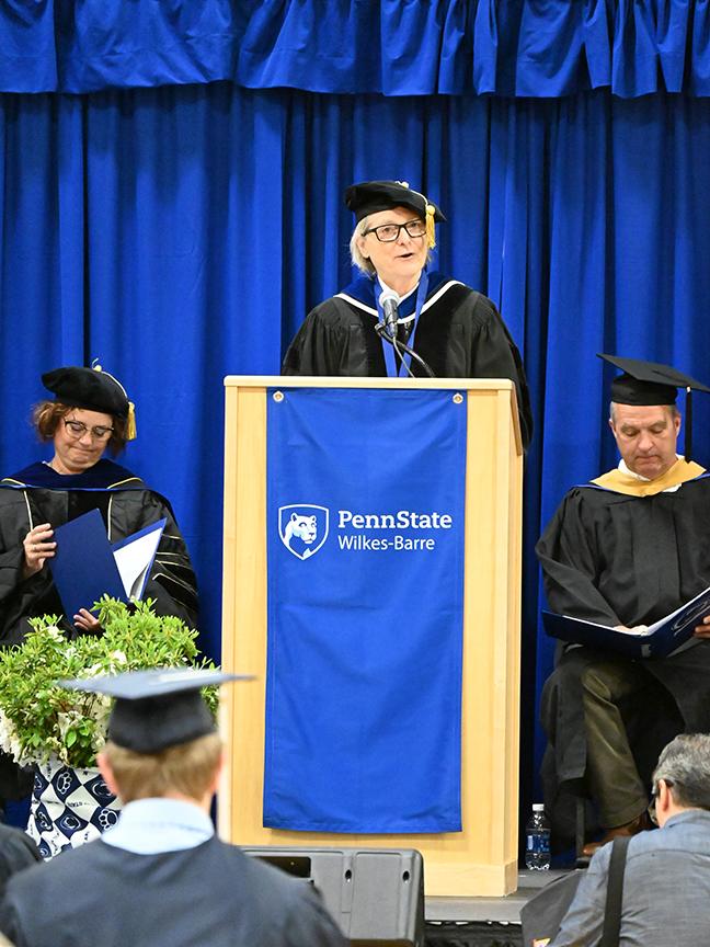 A woman in academic regalia speaks from the podium.
