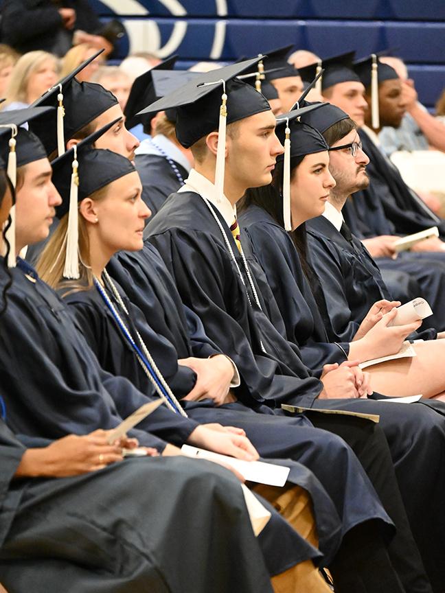Graduates in caps and gowns listen to a speaker.