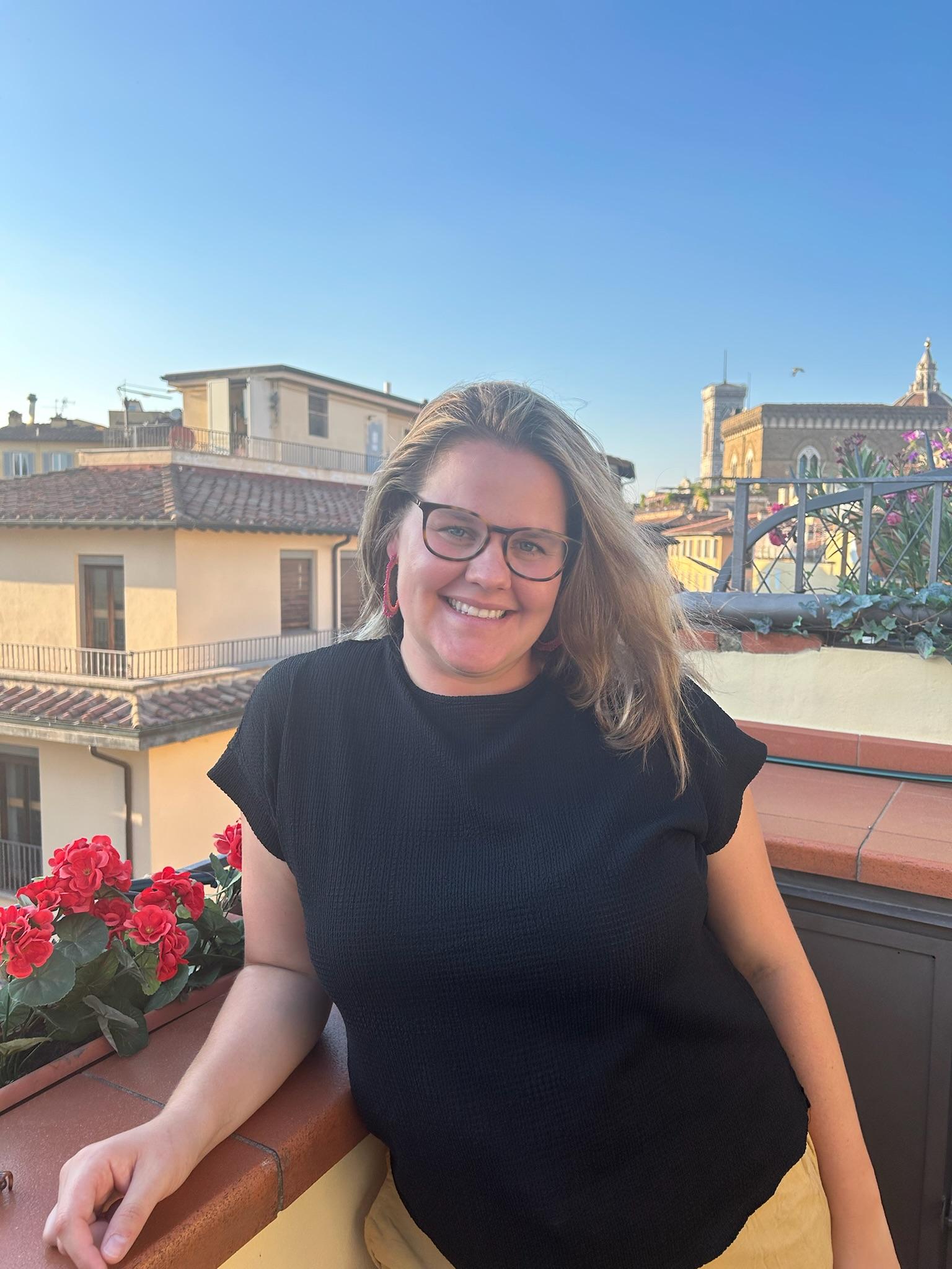 Carly Pourzand smiling while standing on a roof top on a sunny day, with red flowers in a pot and other rooftops in the background..