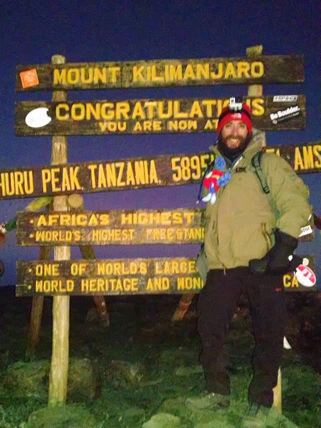 A man in winter gear standing in front of a wooden sign that says "Mount Kilimanjaro."