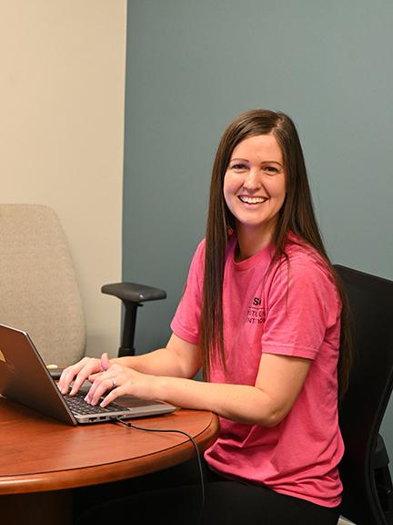 A woman sitting in front of a computer, looking at the camera and smiling.