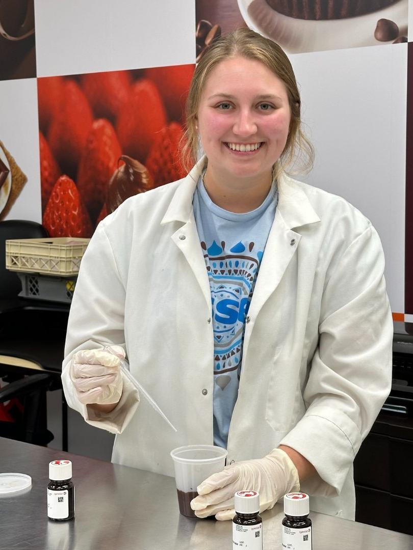 A student wearing a white lab coat holds a beaker and dropper