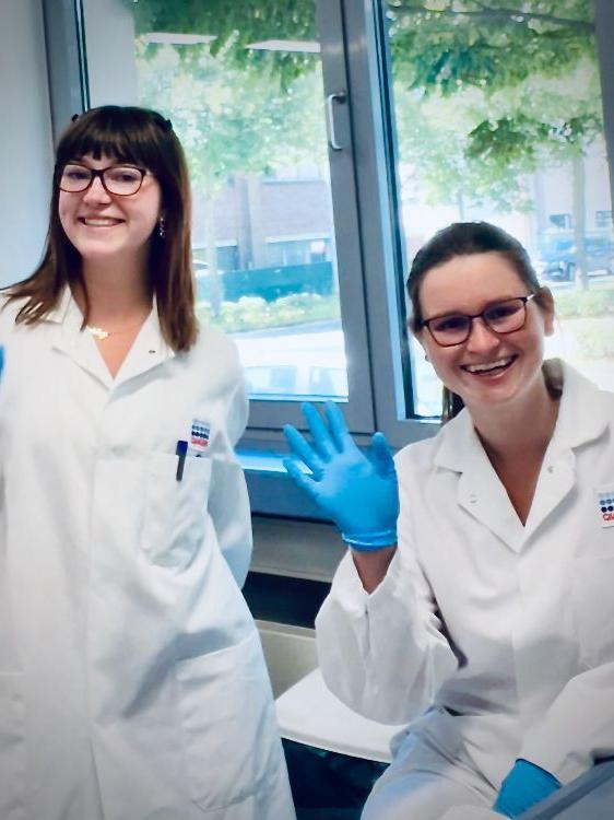 Two students wearing lab coats and blue rubber gloves smile for the camera