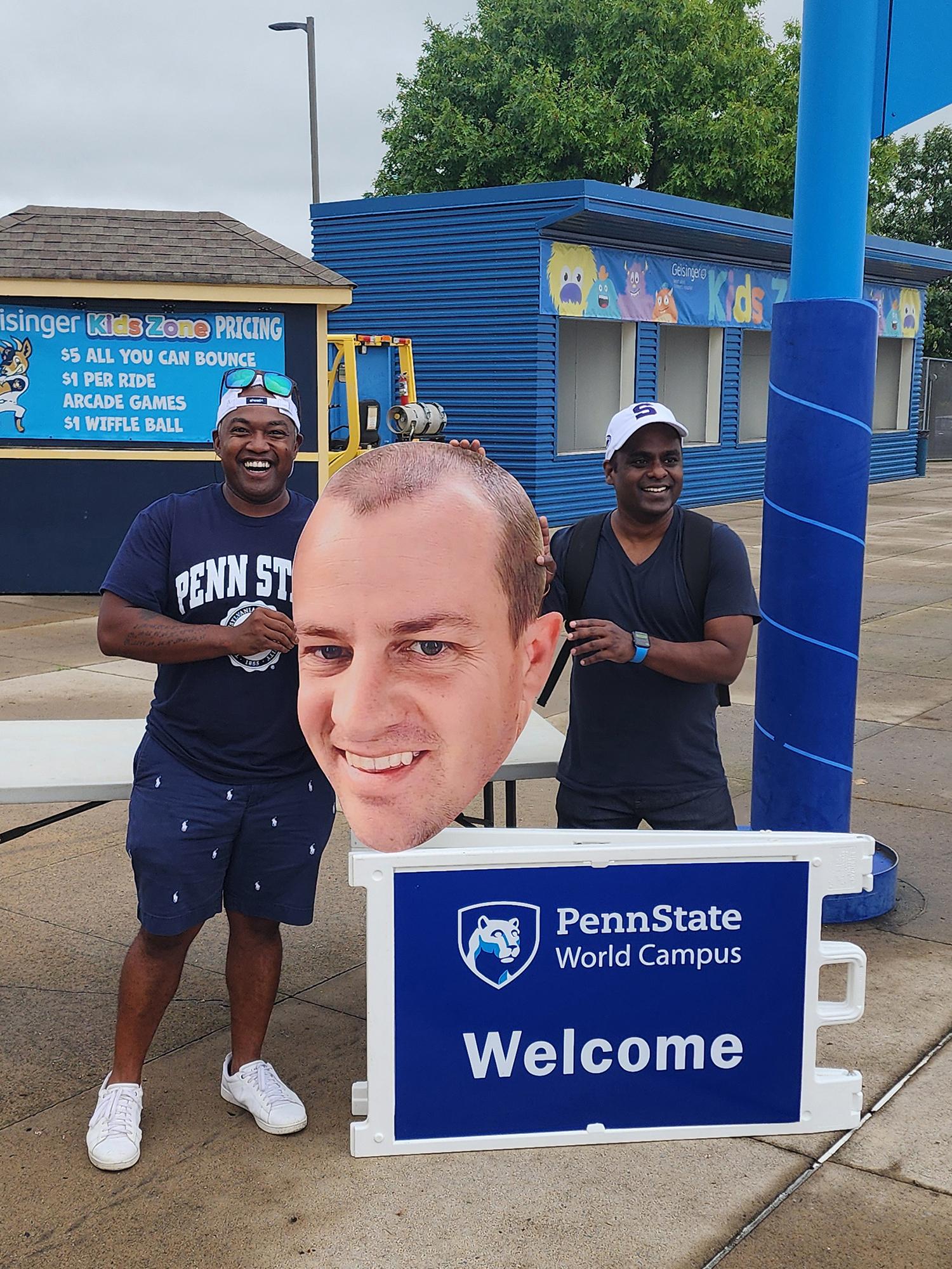 Two men pose in front of a sign with a printed cutout of a man's head.