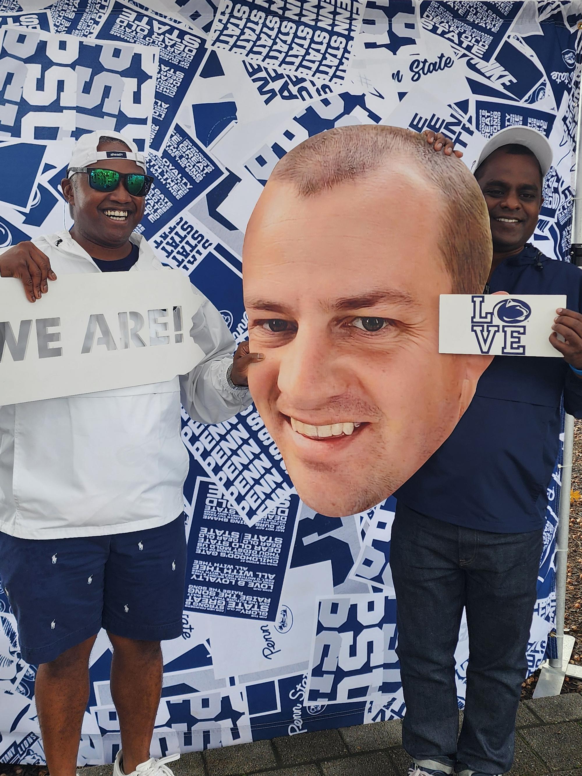 Two men pose with a large cutout of another man's face in front of a blue and white background.