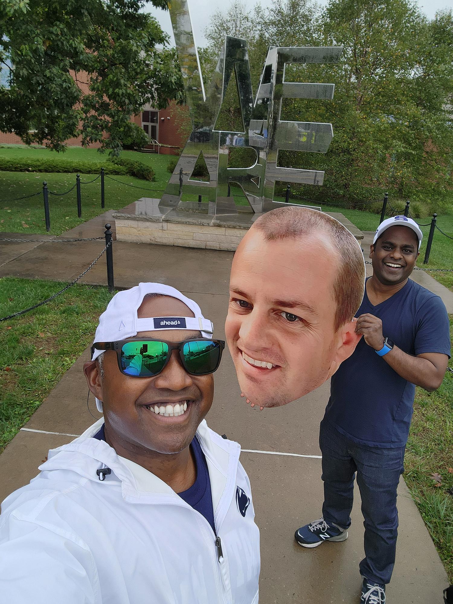 Two men pose in front of the "We Are" sculpture with a printed cutout of a man's head.