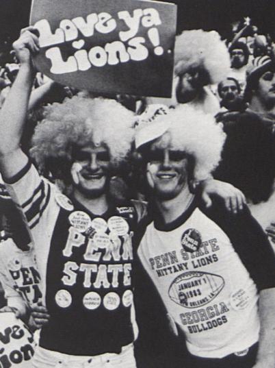 Two football fans wear Penn State gear and hold a sign that reads "Love Ya Lions"
