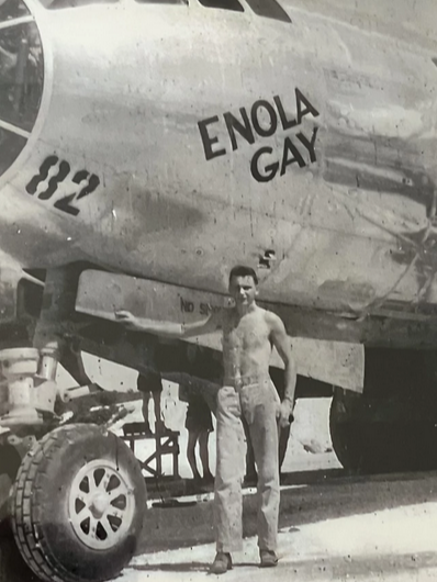 A black-and-white image of a man standing next to a plane with the words "Enola Gay" on its side