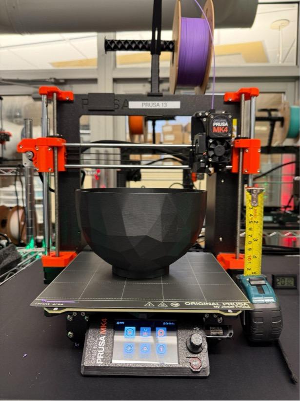 A close-up of a Prusa MK4 3D printer actively printing a large, faceted black bowl. The printer's touchscreen interface is visible at the bottom, displaying various settings. A spool of purple filament feeds into the extruder at the top. To the right of the printer are a measuring tape and a digital caliper to measure the size of the print. In the background are additional 3D printers and filament spools in a well-equipped maker space.