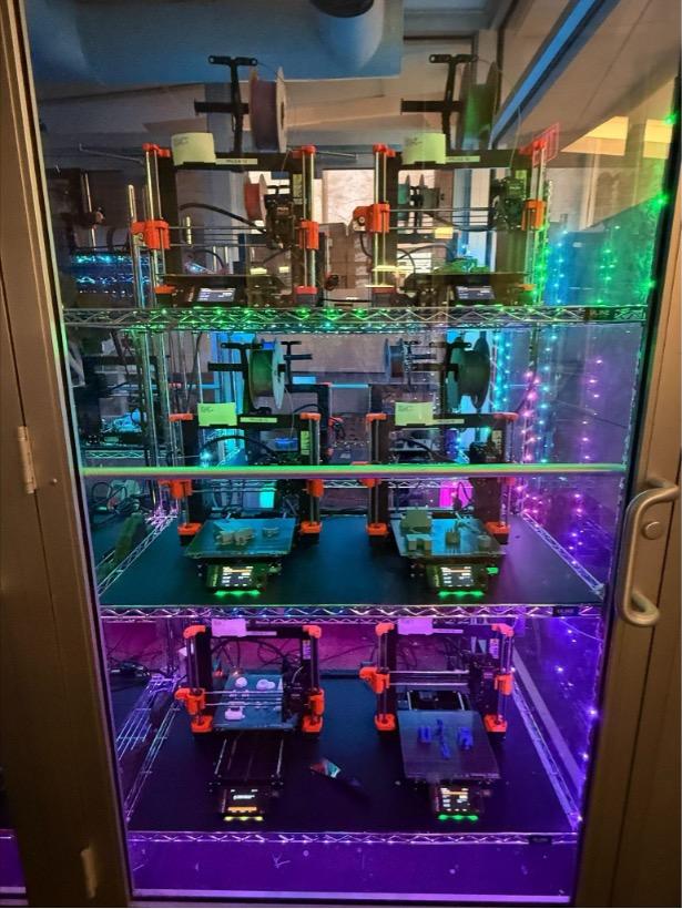 A 3D printing lab featuring multiple Prusa 3D printers arranged on metal shelves in an enclosed glass cabinet. The printers are actively printing various objects, with filament spools mounted above each machine. Green and purple LED lights illuminate the workspace, creating a high-tech, inviting environment and a slight reflection on the glass.
