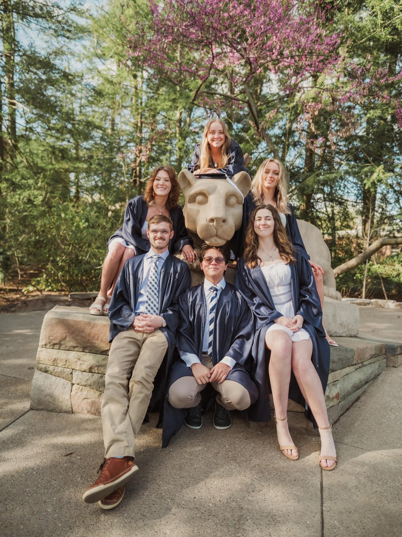 A group of students in graduation robes pose with the Nittany Lion Shrine.