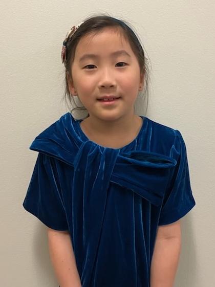 First-grade student wearing a blue dress standing in front of a white backdrop