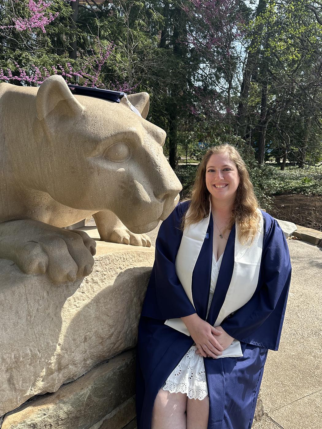 A woman wearing a blue graduation cap and gown sitting next to the Nittany Lion Shrine.