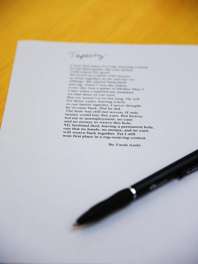 A close-up of a printed poem titled Tapestry by Farah Arabi sits on a wooden table. A black pen rests diagonally across the lower corner of the paper. The poem discusses themes of loss, resilience, and weaving as metaphor, with part of the text clearly legible.