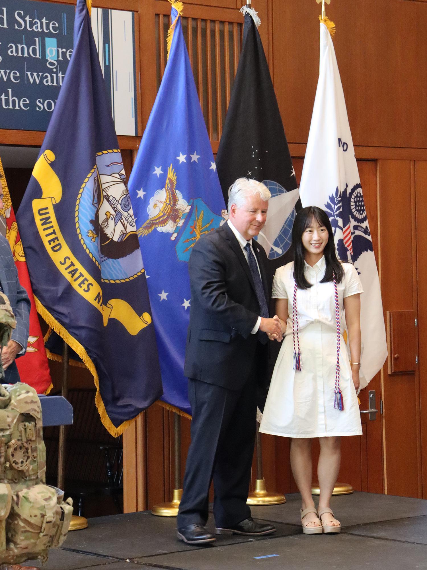 A woman wearing a white dress shakes the hand of a man wearing a suit on a stage.