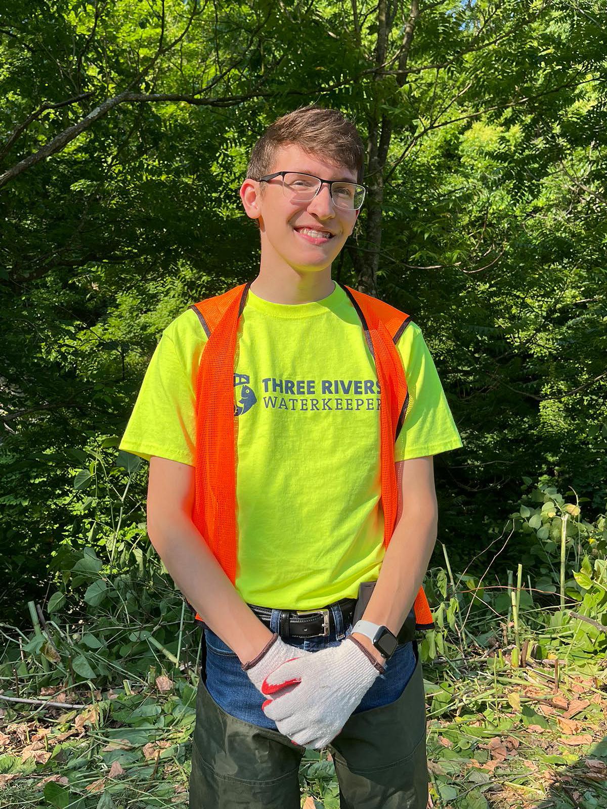 Student in a bright green t-shirt and bright orange vest standing in front of trees